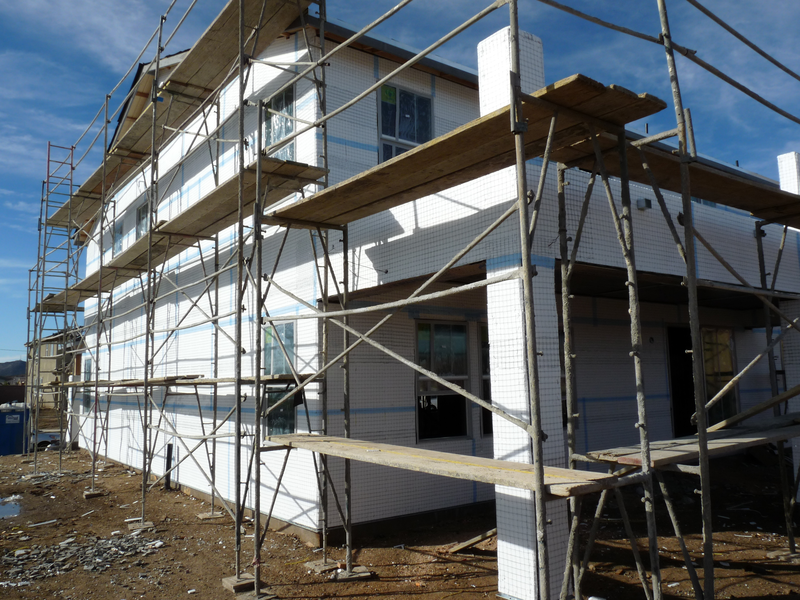 A construction scaffolding against two exterior walls of a house. The walls are wrapped continuously in white rigid insulation boards sealed with blue tape.